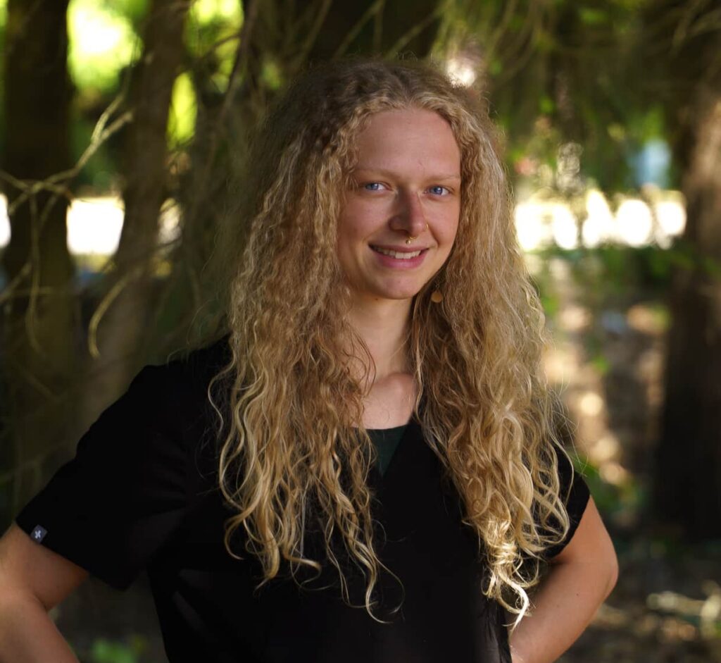 An image of a young blonde woman standing with her hands on her hips and smiling. 