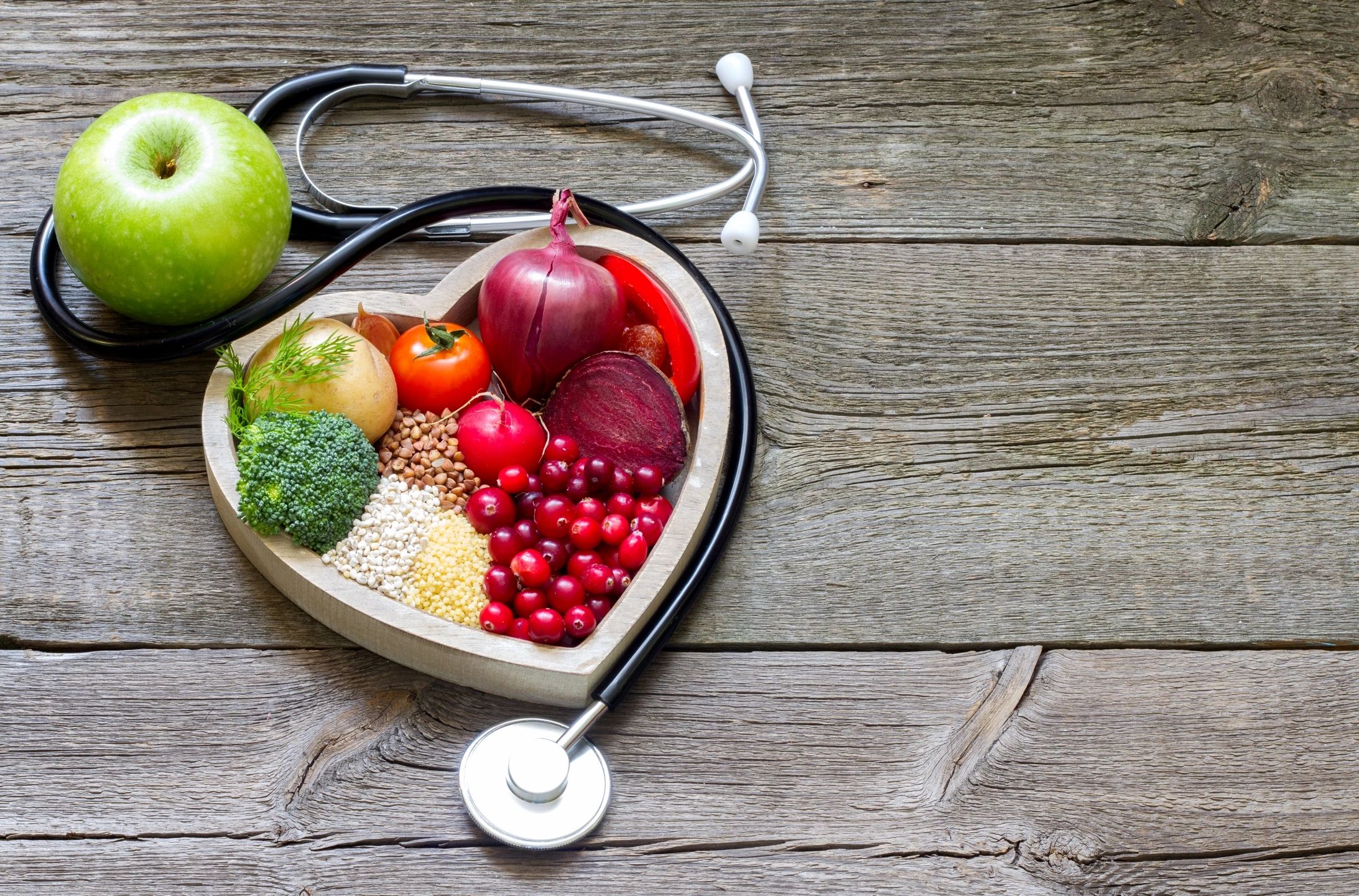 An image of a stethoscope on a wooden table next to an apple and a wooden heart-shaped dish filled with healthy food.
