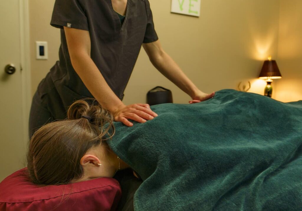 An image of a patient laying face-down on a massage table, covered with a dark green towel, receiving a massage from a female massage therapist.